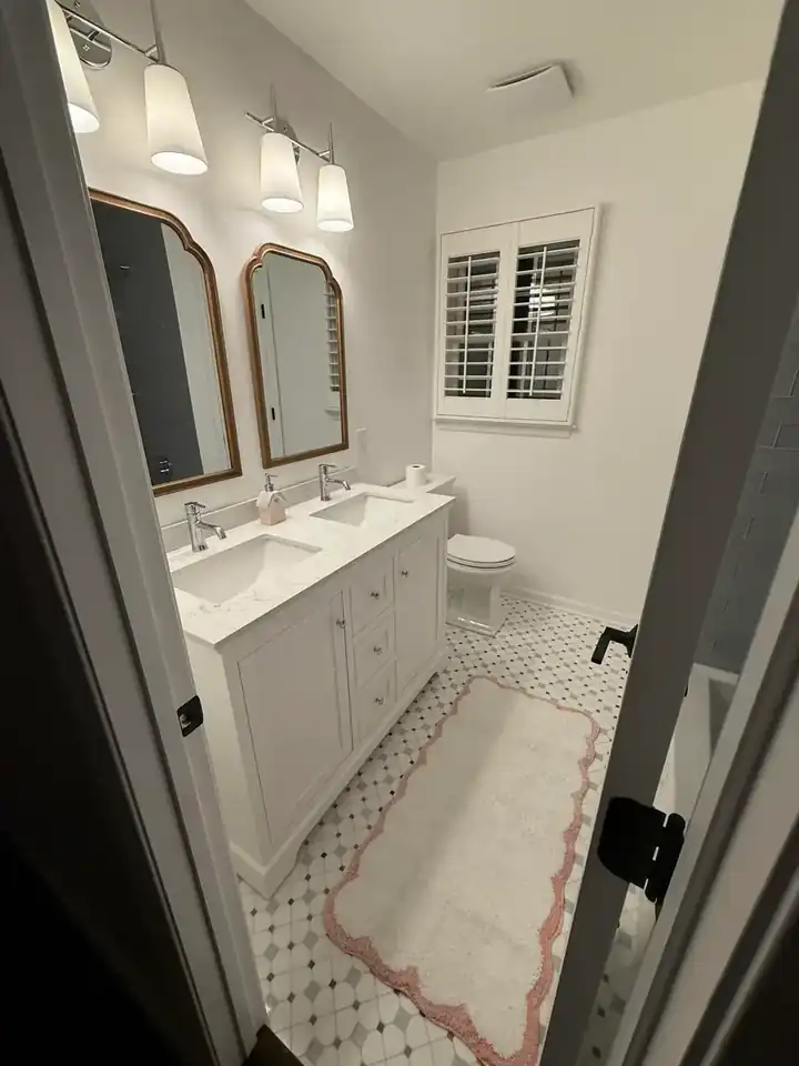 Finished bathroom with white double vanity, twin mirrors, patterned tile floor, window shutters, and coordinated lighting
