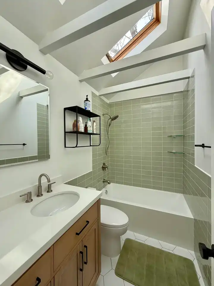 Finished bathroom with vaulted ceiling beams, skylight, green tile tub surround, wood vanity, mirror, and lighting
