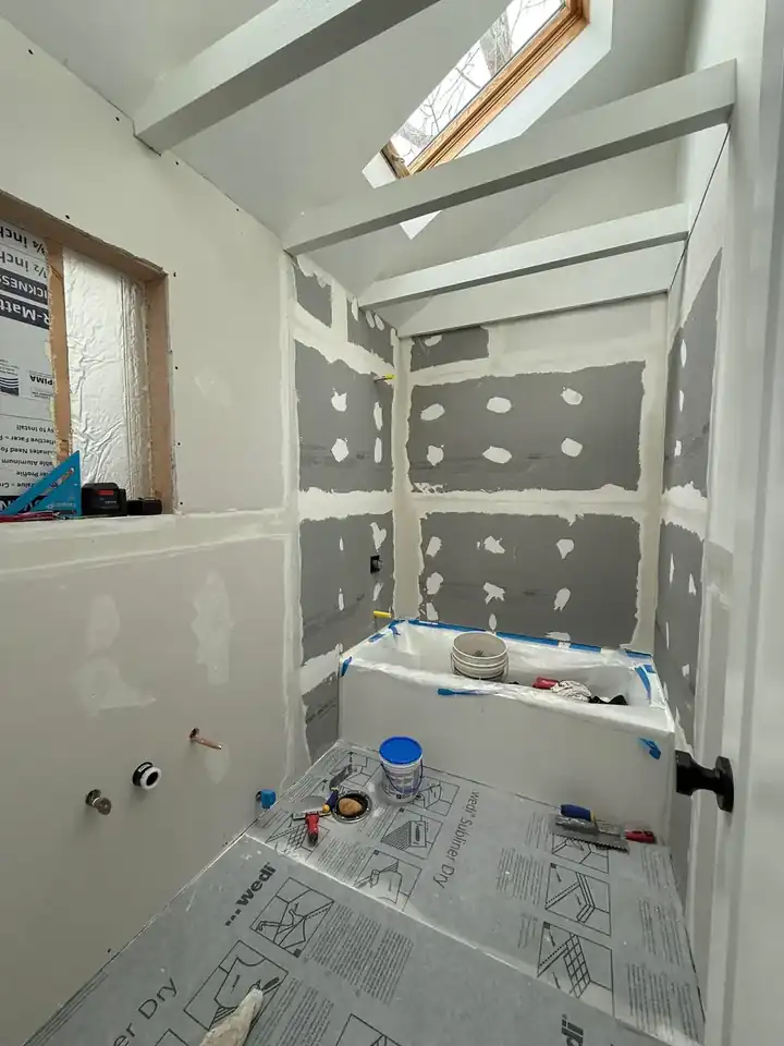 Bathroom renovation progress with wall board, taped seams, protected floor, tub area, vaulted ceiling, and skylight